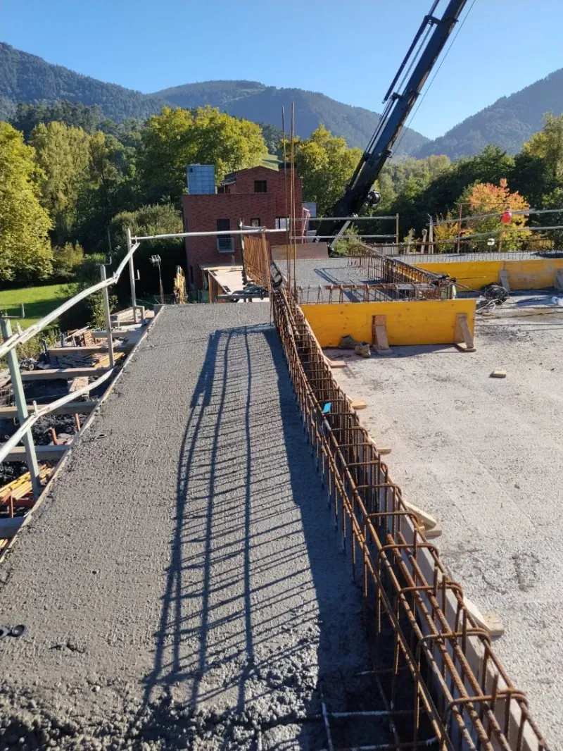 Pasarela de construcción con barras de refuerzo y encofrado de hormigón junto a una grúa, montañas al fondo.