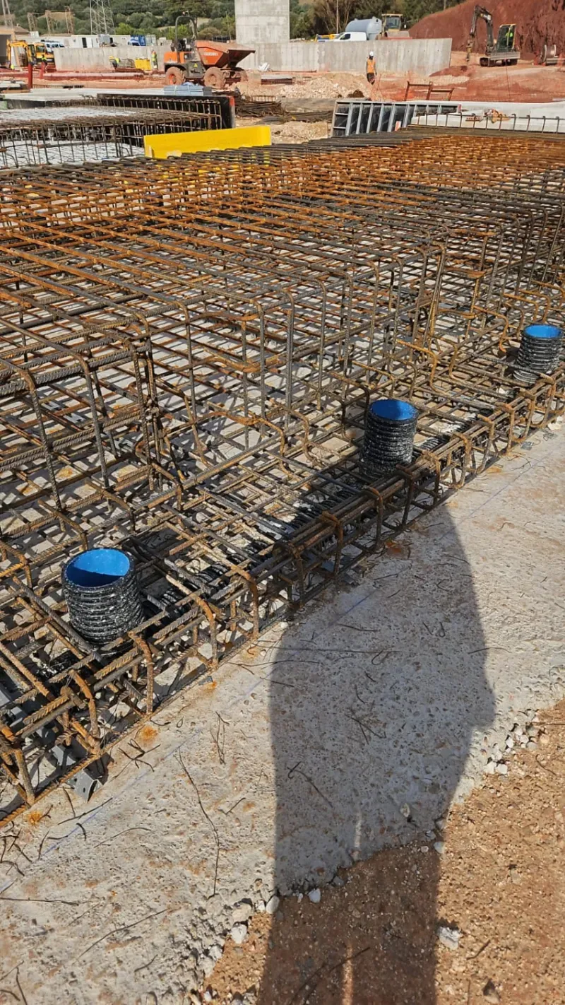 Rejilla de barras de refuerzo en una obra de construcción para una cimentación de hormigón con tapas de tuberías azules y la sombra de un trabajador en primer plano.