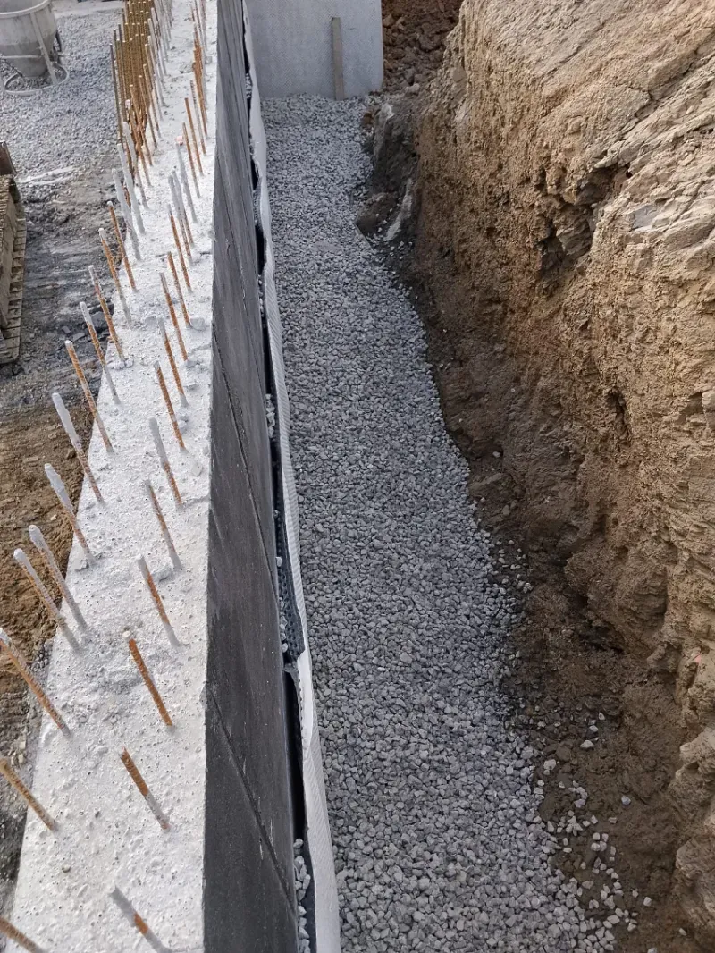 Zanja estrecha de grava junto a un muro de tierra, con una hilera de estacas metálicas expuestas a lo largo del borde izquierdo.