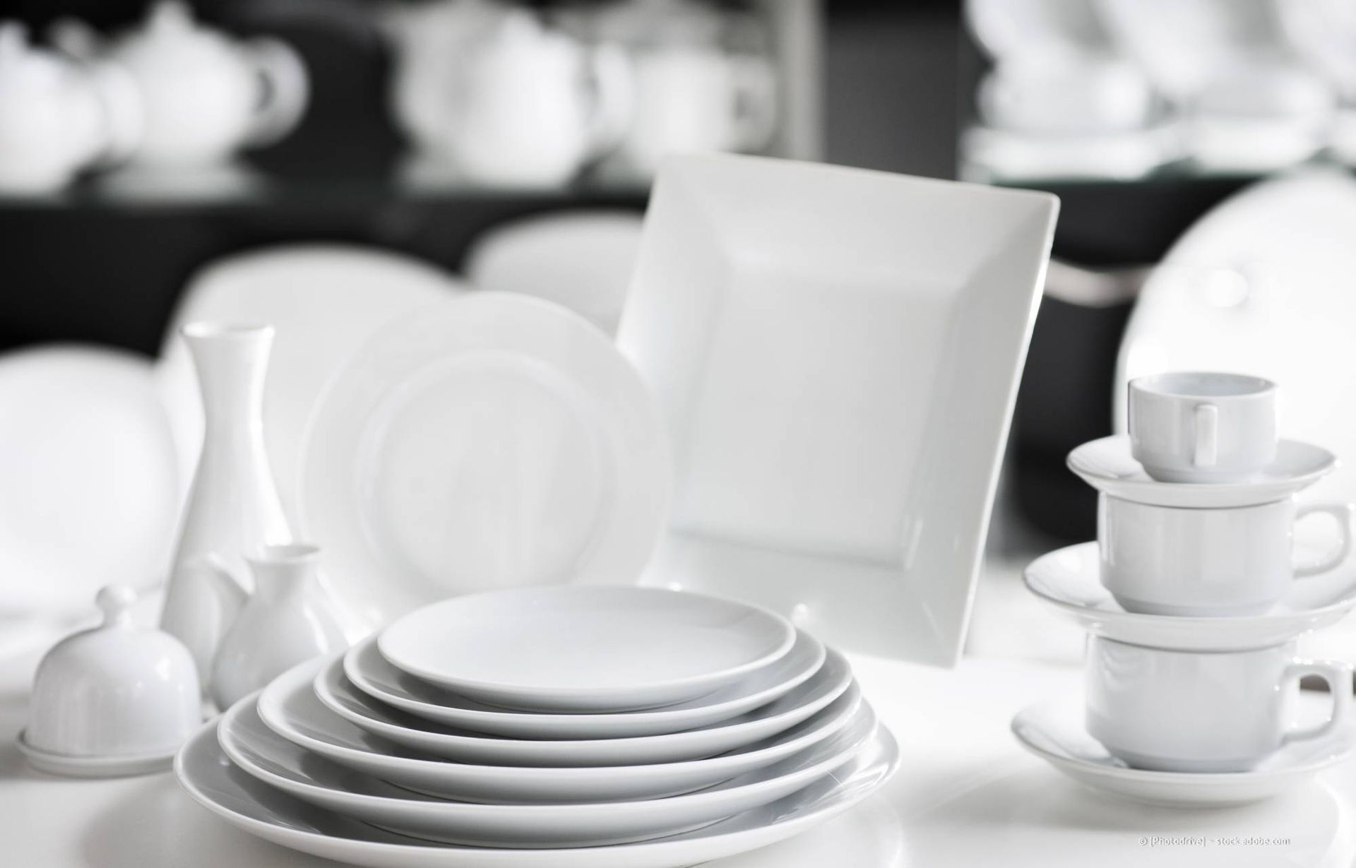 Ein Stapel weißer Teller und Tassen auf einer hellen Fläche vor einem Regal mit ähnlichen Gegenständen.