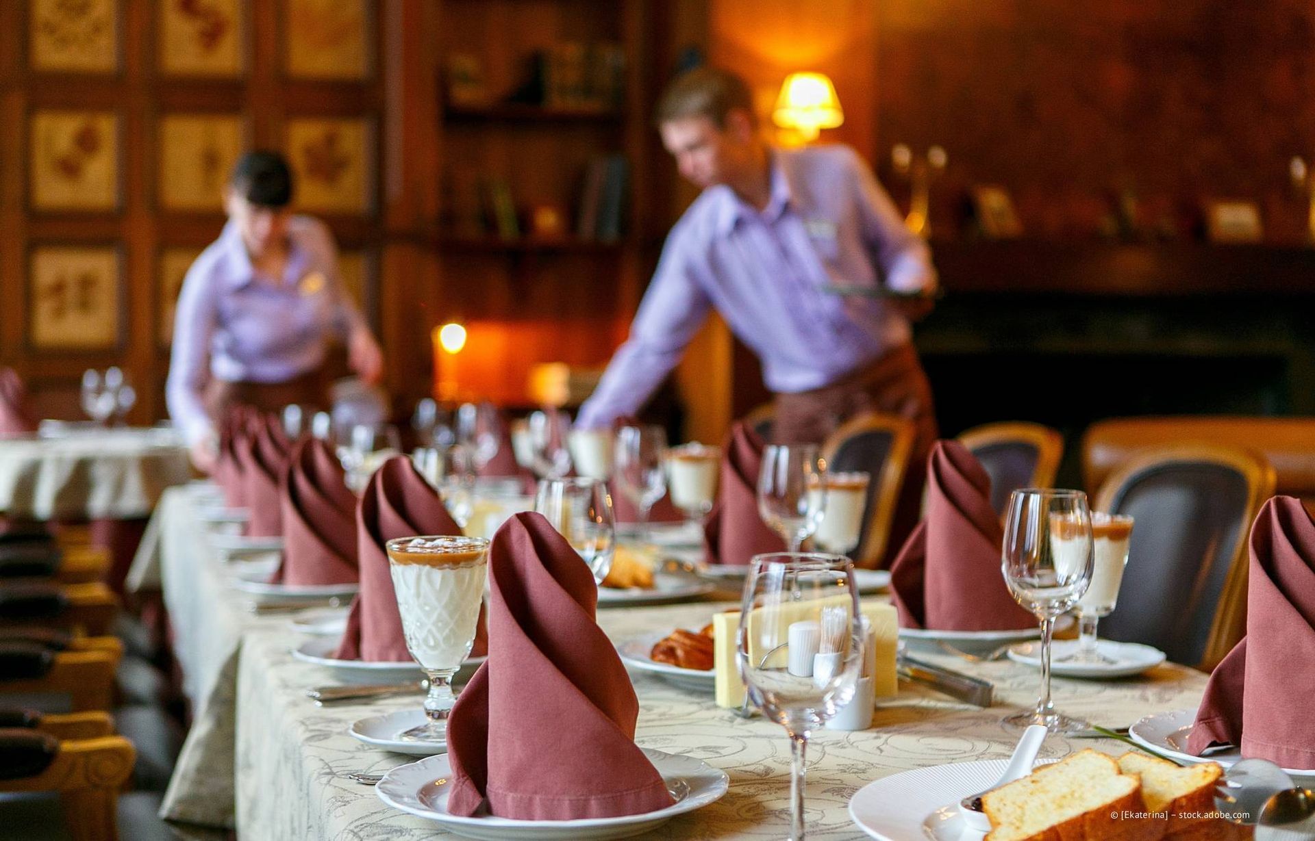 Ein Restauranttisch ist mit Servietten, Gläsern und Speisen gedeckt, das Personal ist mit dem Arrangieren beschäftigt.