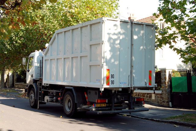 Camion-poubelle blanc dans une rue bordée d'arbres, vu de l'arrière.