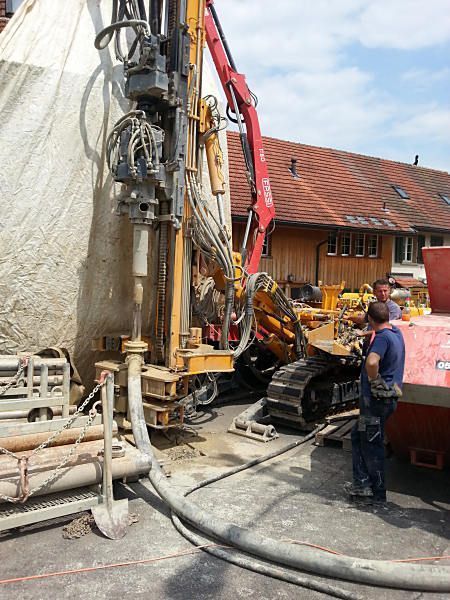 Bohranlage neben einem Haus, mit einem Arbeiter und einem Bagger auf einer Baustelle.
