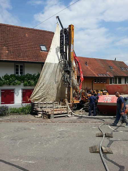 Arbeiter bedienen eine grosse Bohranlage neben einem Haus; davor befinden sich eine mit einer Plane abgedeckte Konstruktion und Schläuche.