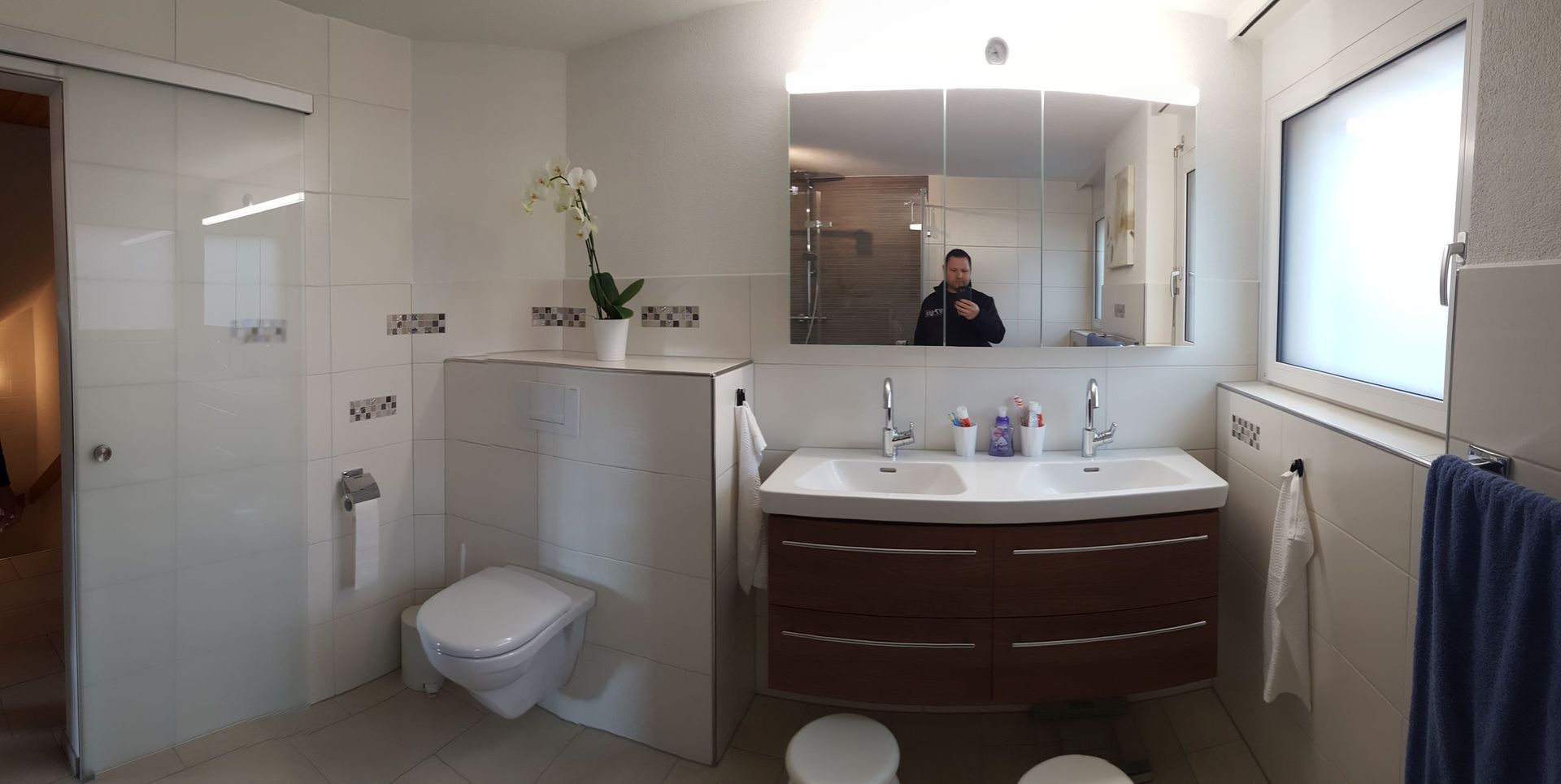 Helles Badezimmer mit Waschbecken, Toilette, Glasdusche, Handtuchhalter und einer Person, die sich im Spiegel spiegelt