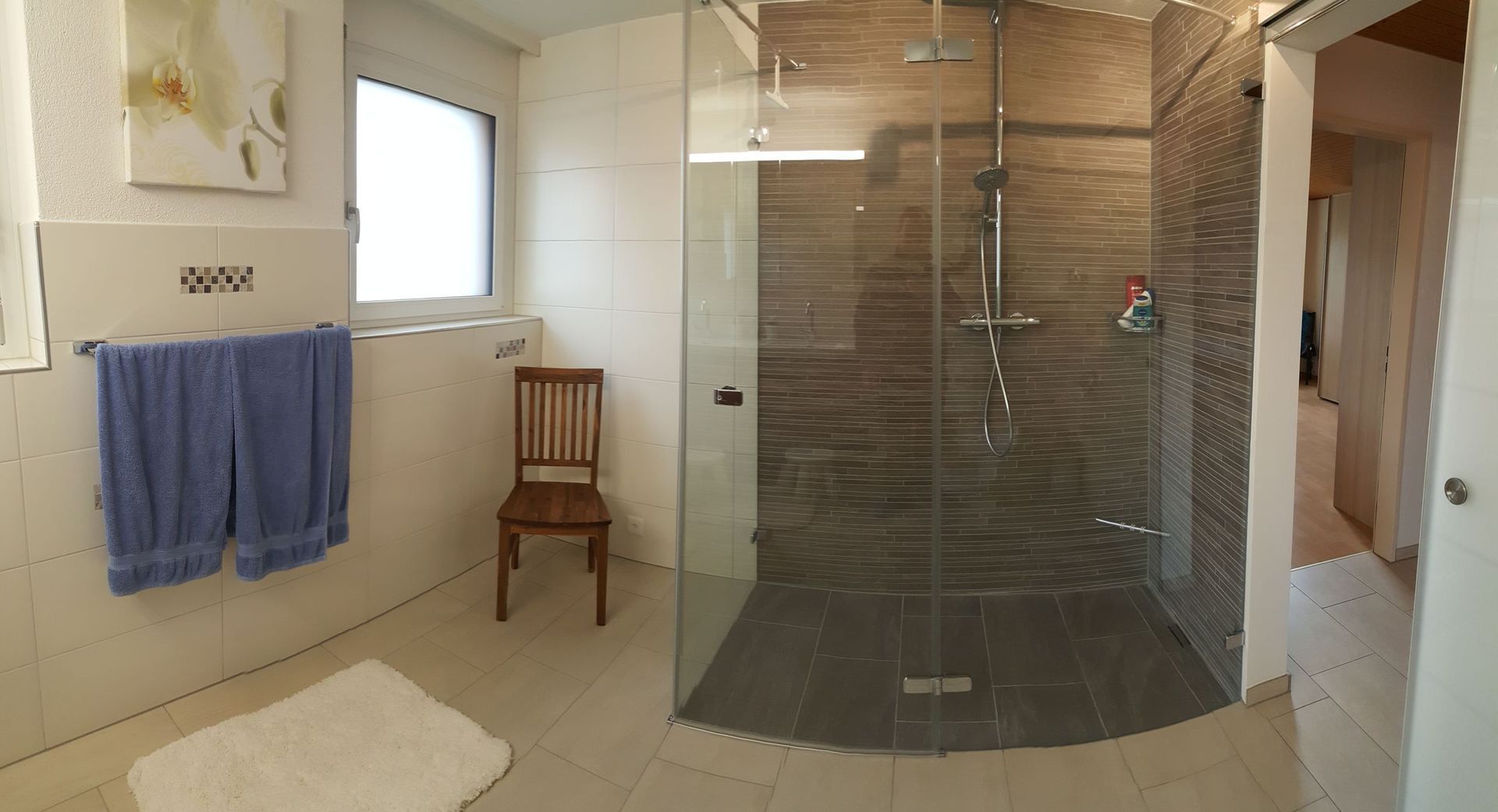 Modernes Badezimmer mit Glasdusche, weissen Fliesen, Holzstuhl, Handtuchhalter und Fenster.
