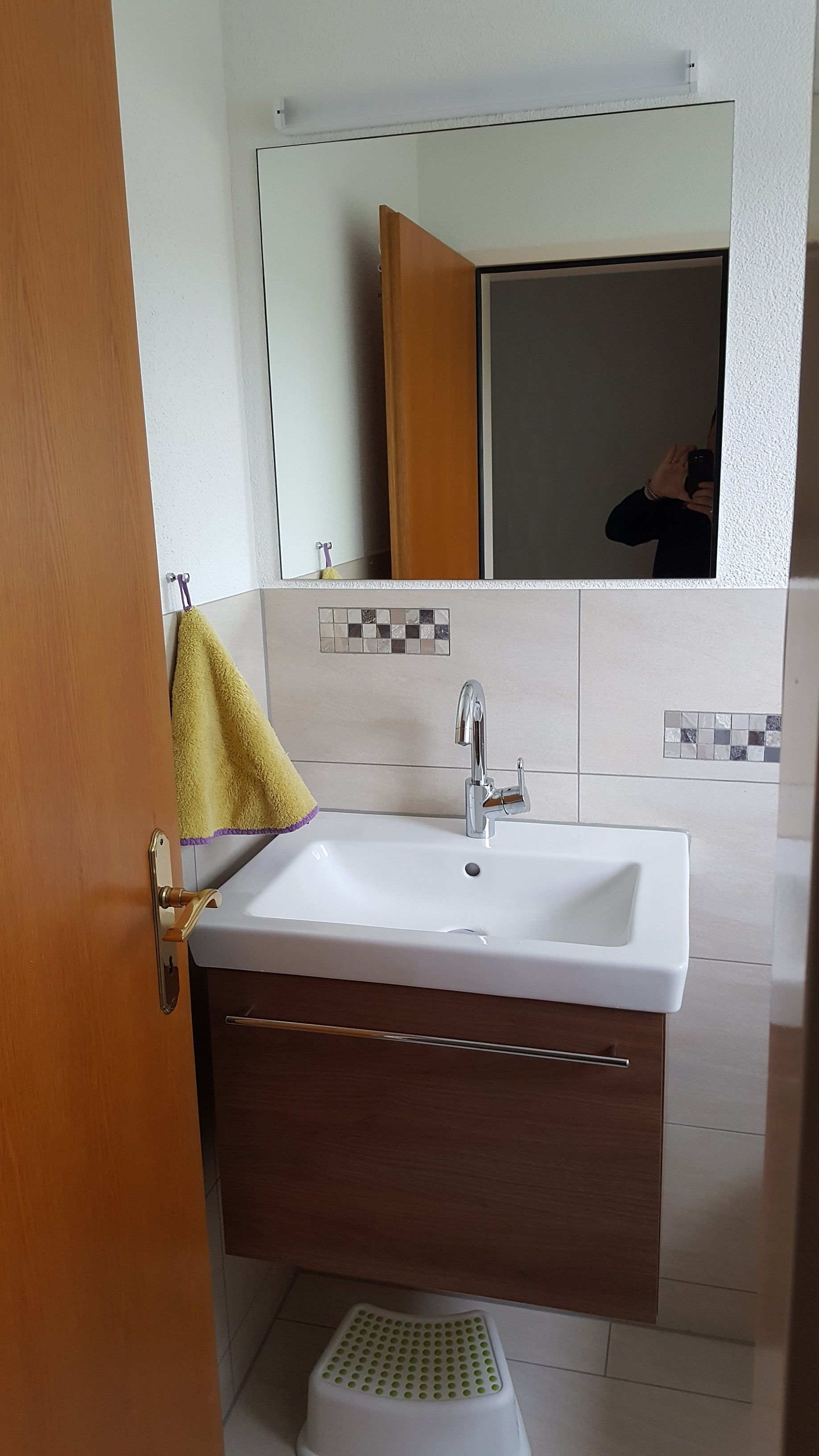 Kleines Badezimmerwaschbecken mit Spiegel, Holzschrank, offener Tür und einem neben dem Waschtisch hängenden Handtuch.