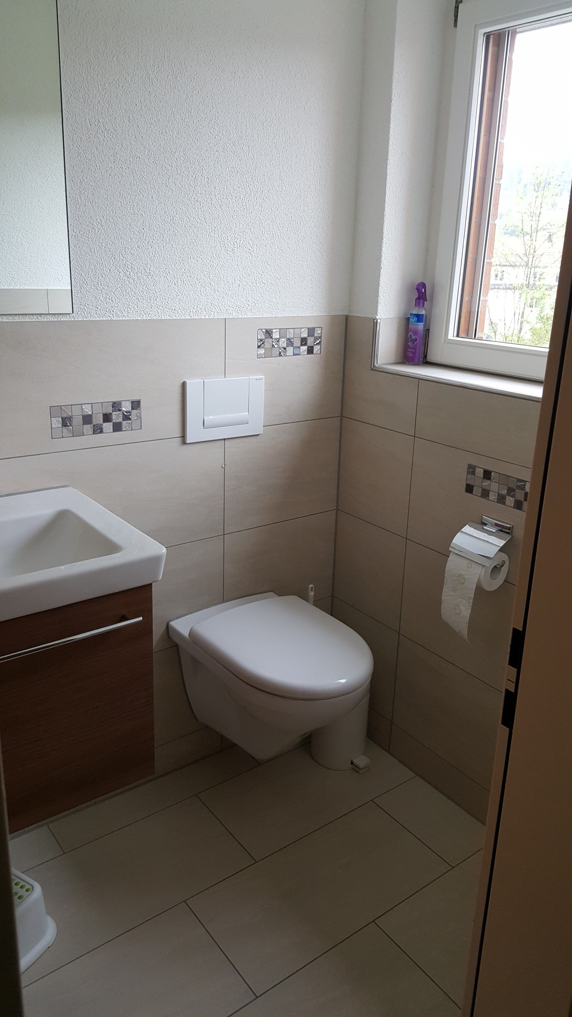 Kleines beigefarbenes Badezimmer mit Toilette, Waschbecken, gefliesten Wänden und Fenster in der Nähe des Toilettenpapierhalters.
