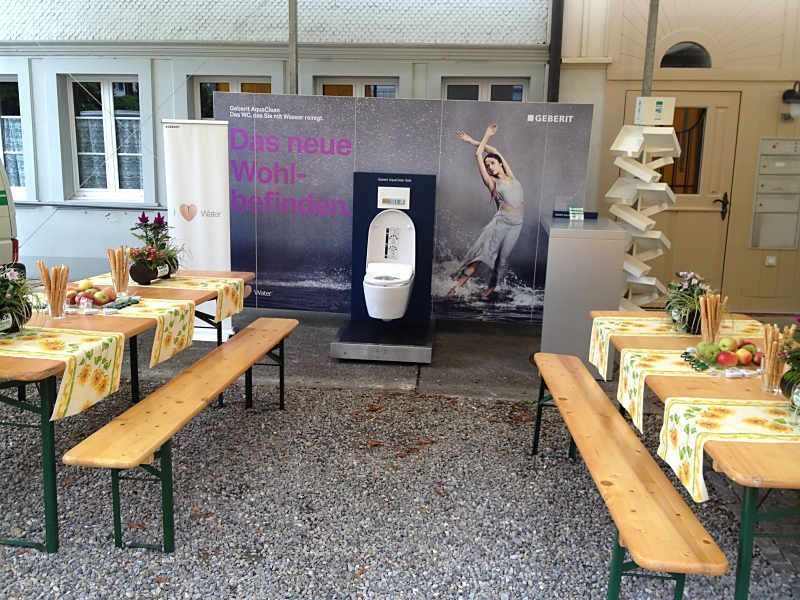 Im Hintergrund sieht man Sitzgelegenheiten im Biergarten mit Bänken und einem Werbebanner für einen Getränkeautomaten.