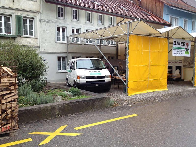 Ein weisser Lieferwagen parkt neben einem Gebäude unter einem gelben Baugerüst auf einer Strasse mit aufgemalten Markierungen.