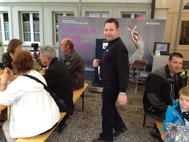Bei einer Indoor-Veranstaltung sitzen und stehen Menschen vor einem Hintergrundplakat; an Tischen unterhalten sich Menschen.
