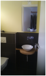 Kleines Badezimmer mit wandmontiertem Waschbecken, Spiegel, schwarzen Fliesen und Toilette auf der linken Seite.