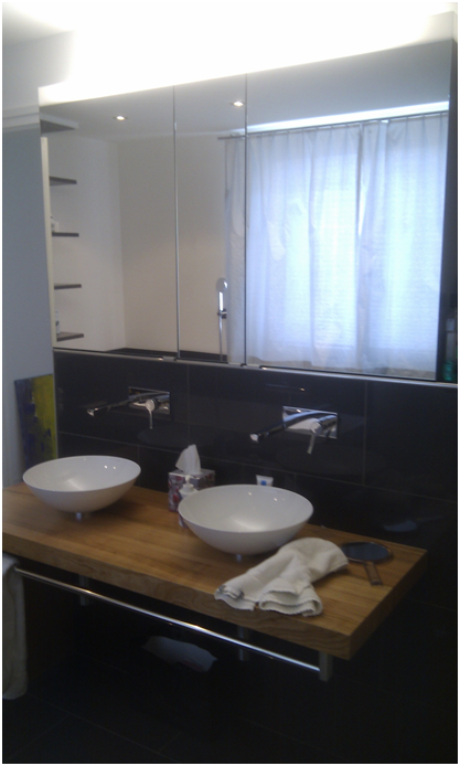 Modernes Badezimmer mit Doppelwaschbecken, Wandspiegel, schwarzen Fliesen und einem Fenster mit transparenten Vorhängen.