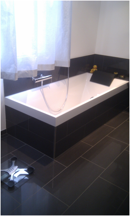 Modernes Badezimmer mit weisser Badewanne, schwarzem Fliesenboden und Fenster mit transparenten Vorhängen.