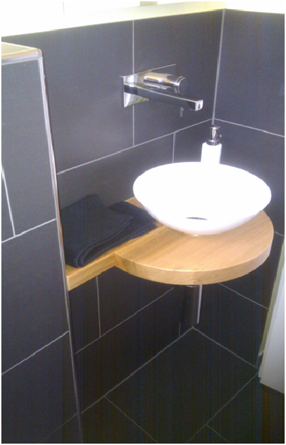 Kleines Badezimmerwaschbecken mit hölzernem Eckregal und schwarzen Fliesen unter einem an der Wand montierten Wasserhahn.