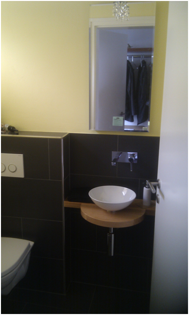 Kleines Badezimmerwaschbecken mit rundem Becken, Spiegel, dunklen Fliesen und gelben Wänden.