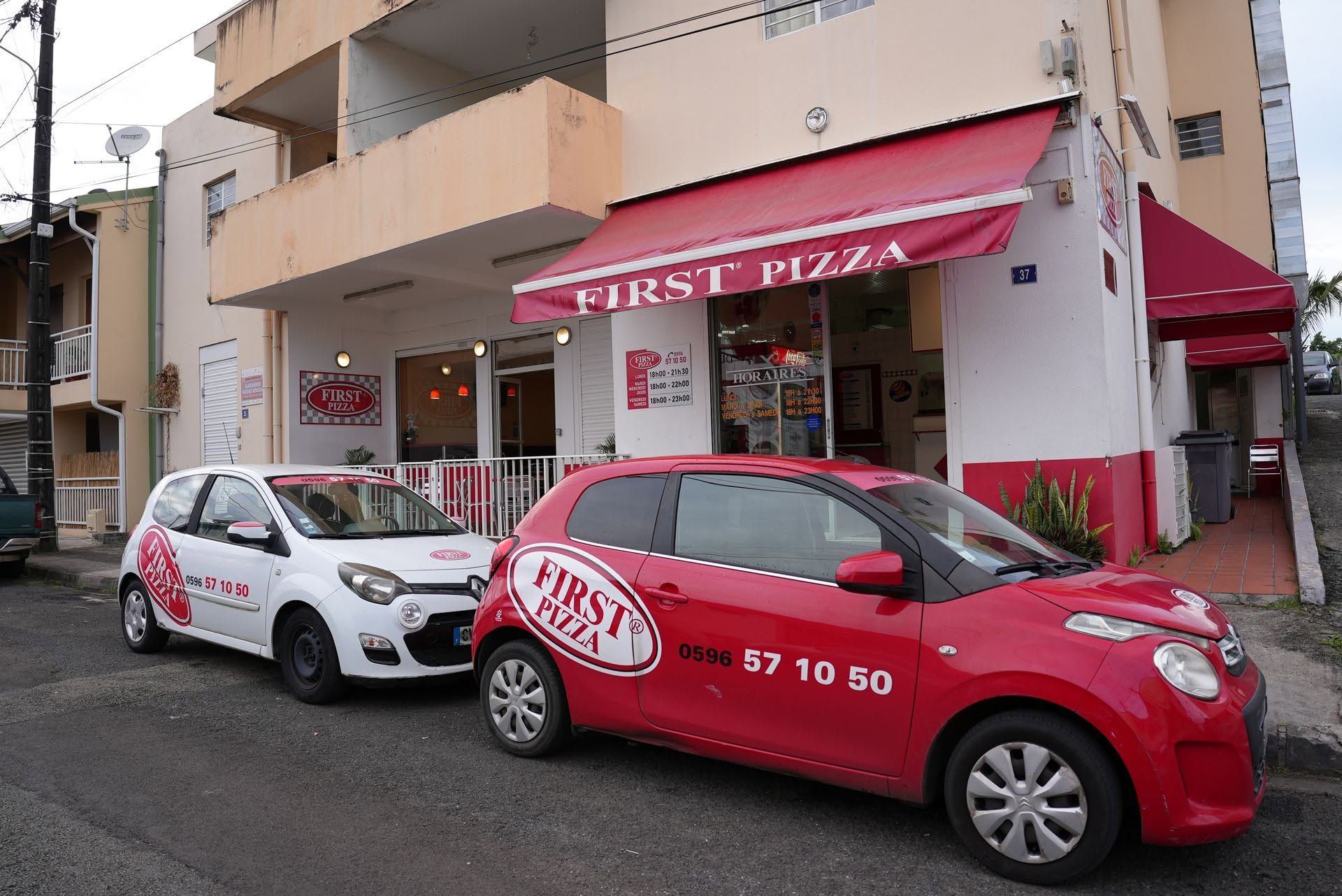 Vue de rue d'une voiture rouge et d'une voiture blanche garées devant le restaurant First Pizza, qui possède un auvent rouge.
