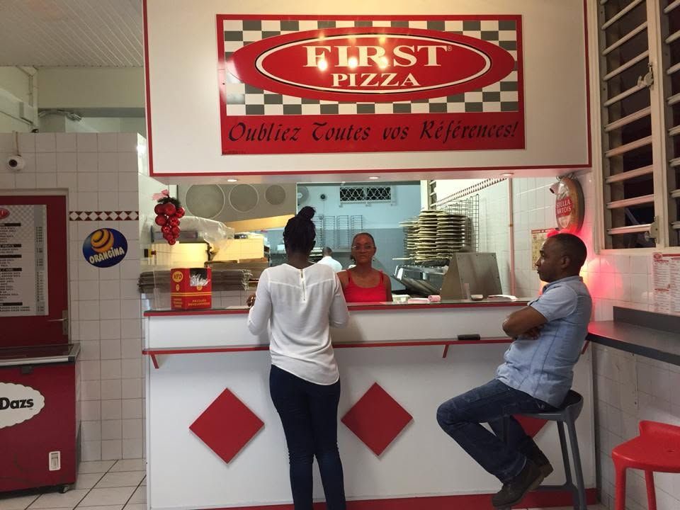 Comptoir à pizzas avec deux clients qui attendent au comptoir d'un restaurant « First Pizza » rouge et blanc