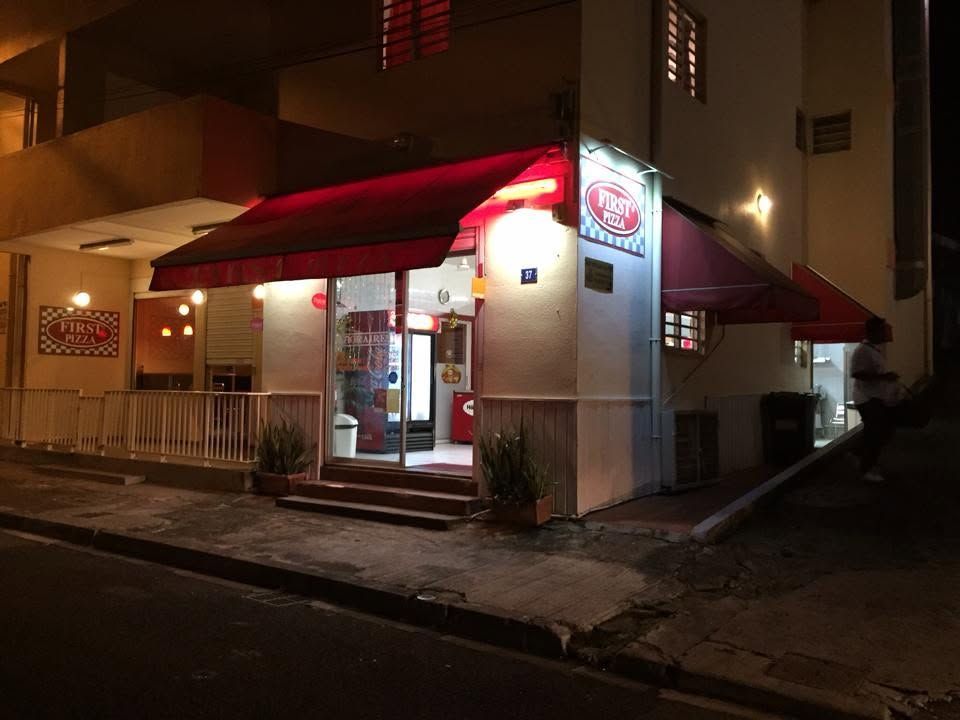 Vue nocturne d'une petite devanture de magasin avec des auvents rouges et une enseigne lumineuse au coin de la rue.