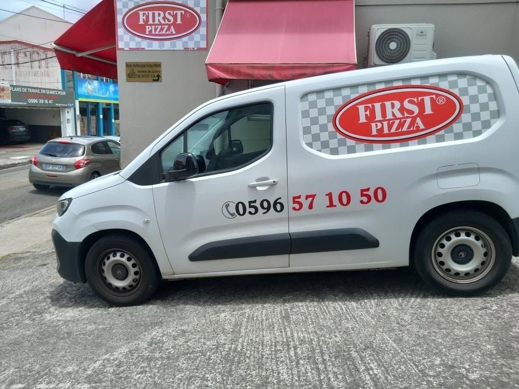 Camionnette blanche de livraison First Pizza garée devant une pizzeria aux auvents rouges.