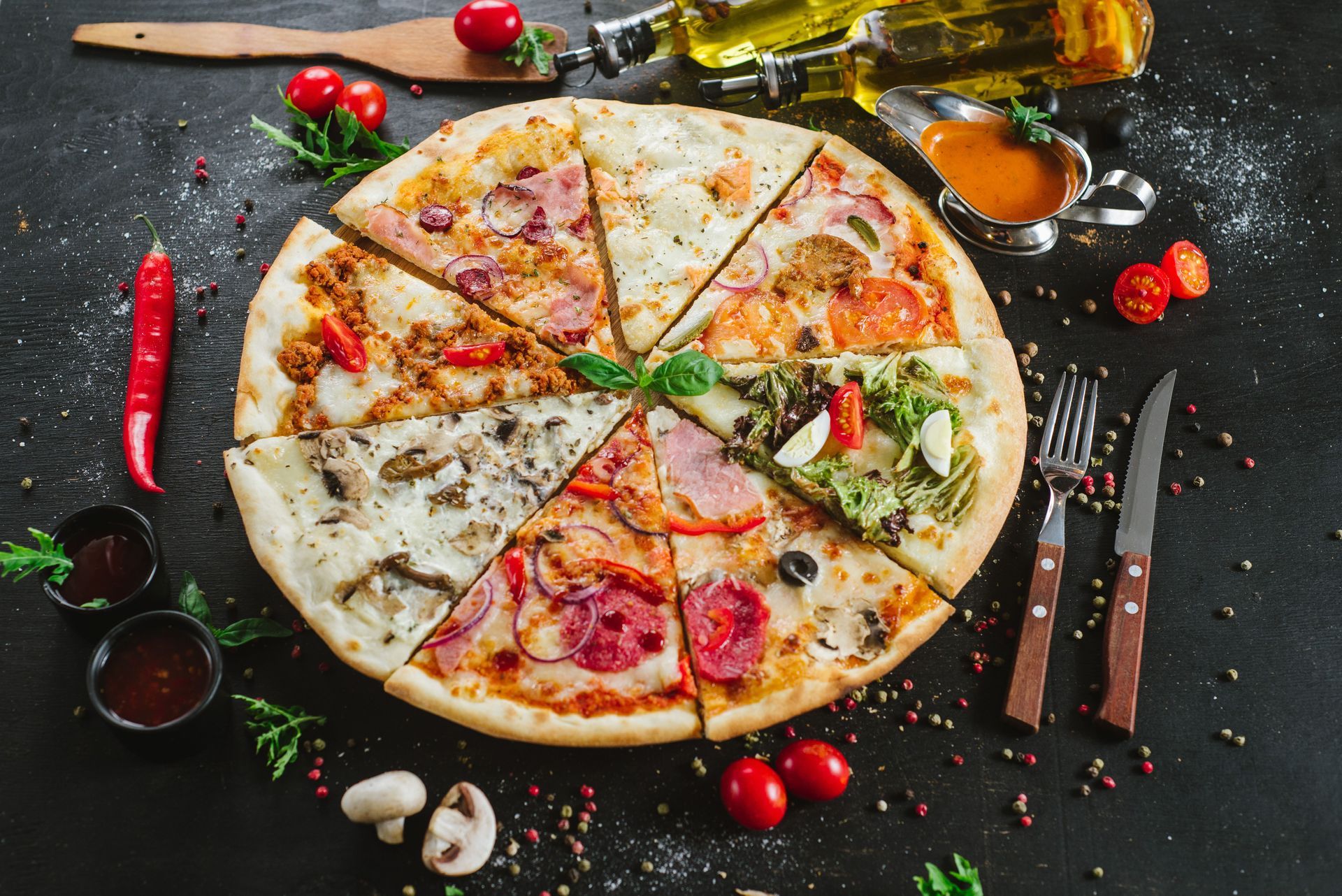 Une pizza sur une table sombre, avec des tomates, des herbes, de l'huile, et une fourchette et un couteau disposés autour.