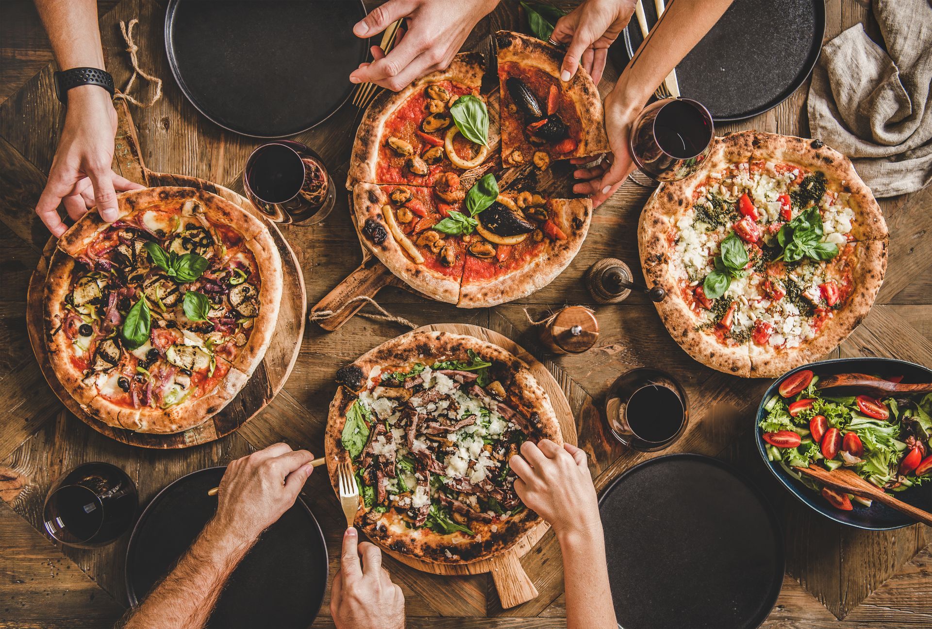 Des gens partagent plusieurs pizzas et salades autour d'une table, les mains venant de tous les côtés.
