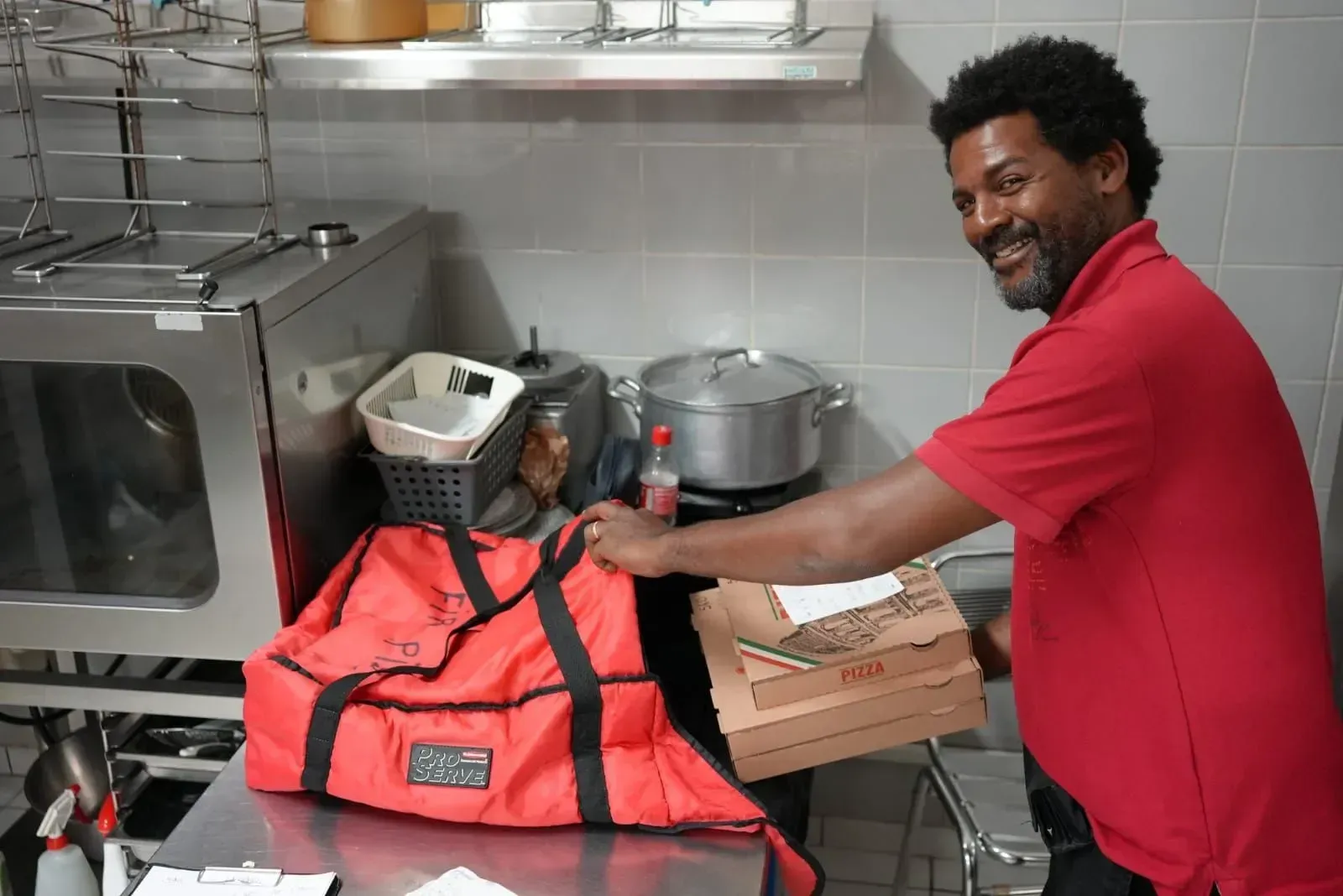 Un homme en chemise rouge décharge un sac de livraison de pizzas dans une cuisine, à côté d'appareils électroménagers en métal.