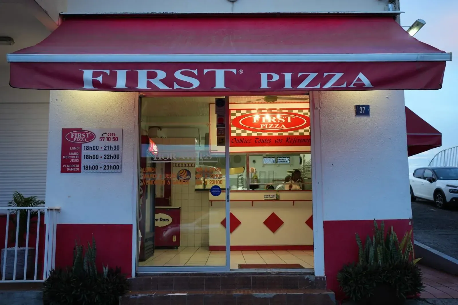 Façade du restaurant First Pizza avec son auvent rouge et sa fenêtre de menu éclairée au crépuscule