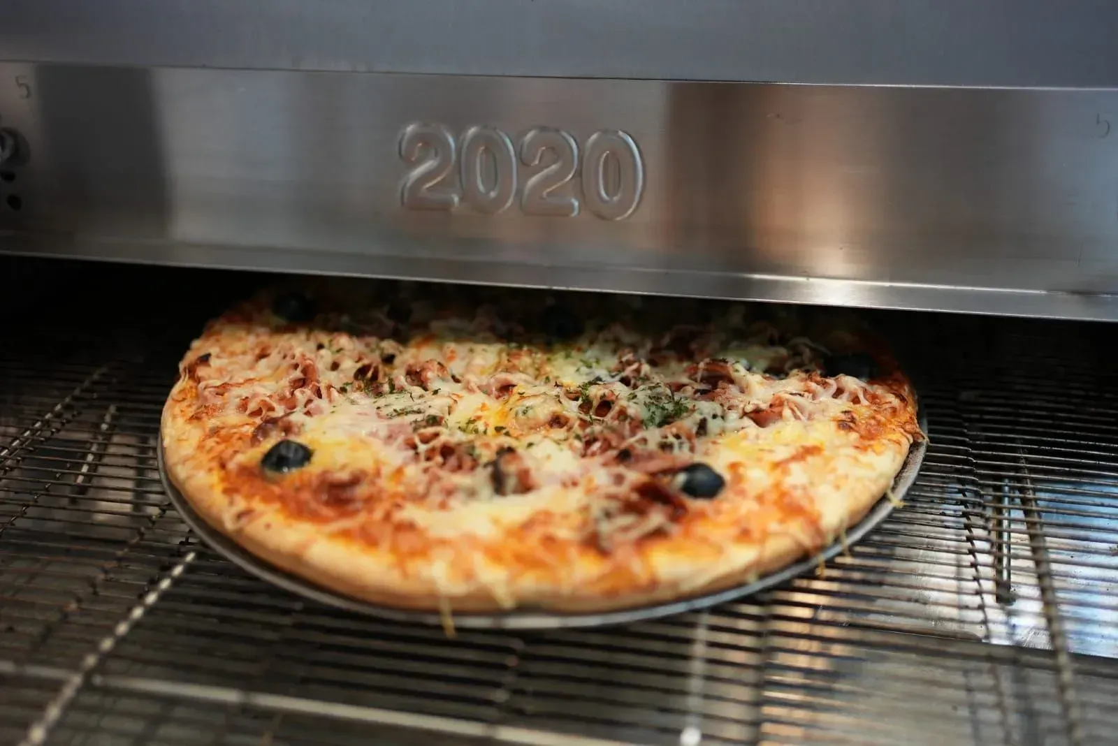 Pizza sur une grille métallique sous un four professionnel, avec du fromage fondu et des garnitures foncées.