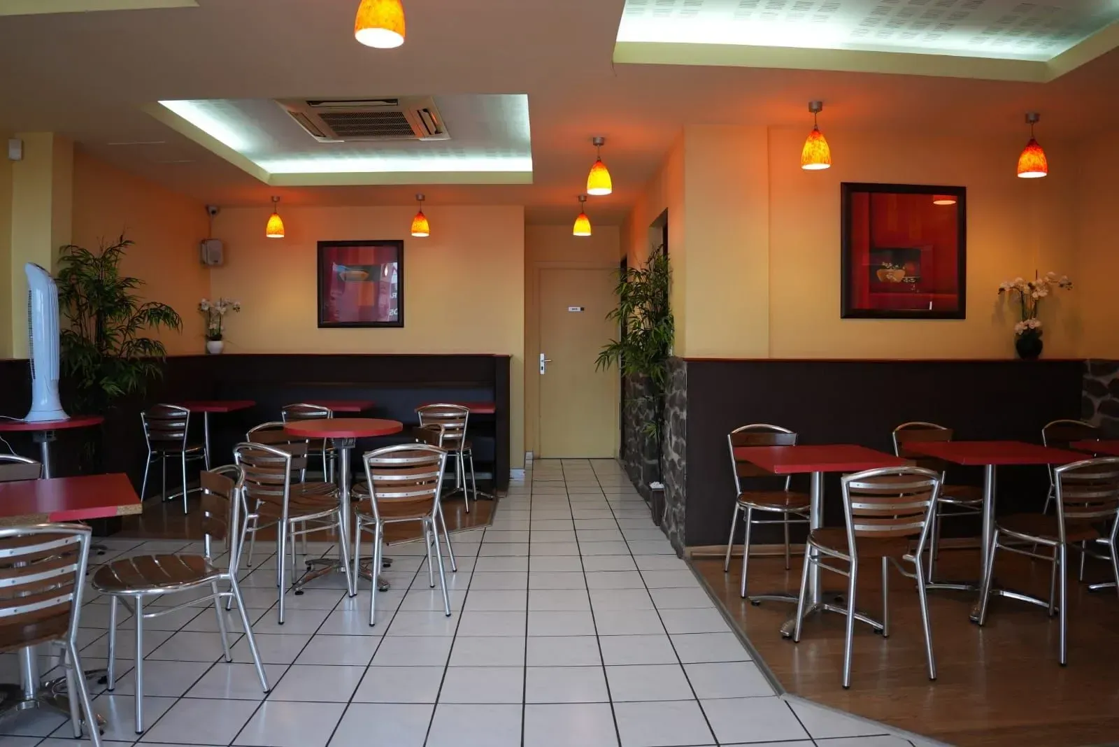 Intérieur d'un café vide avec des tables rouges, des chaises en métal, un sol carrelé et des suspensions lumineuses à l'ambiance chaleureuse.