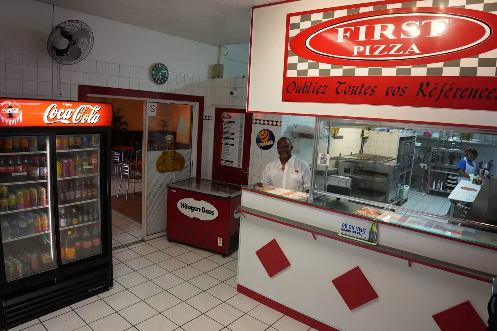 Intérieur d'une pizzeria avec un réfrigérateur et un comptoir rouge et blanc.