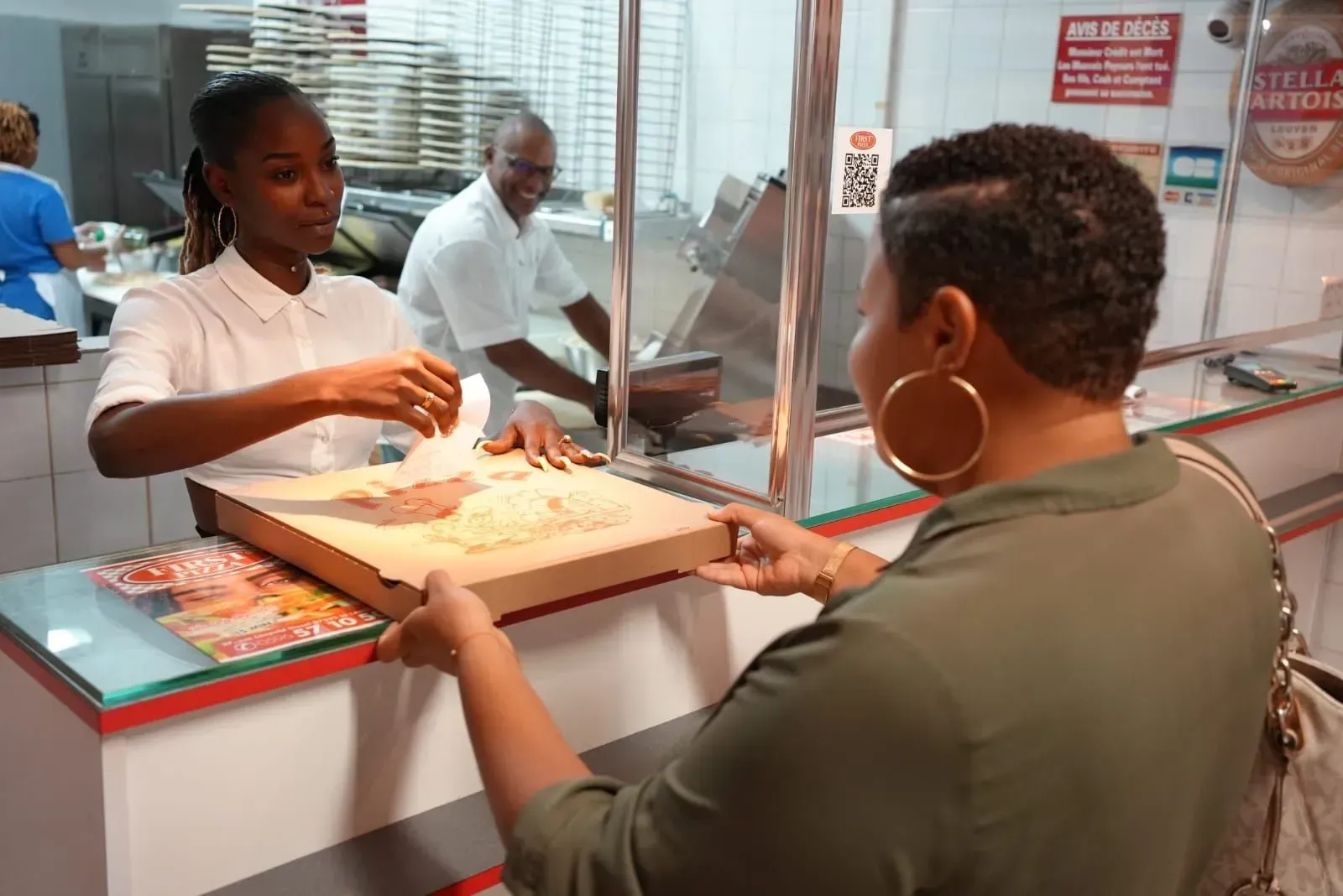 Client recevant une pizza en boîte au comptoir d'une pizzeria à travers une vitre en plexiglas