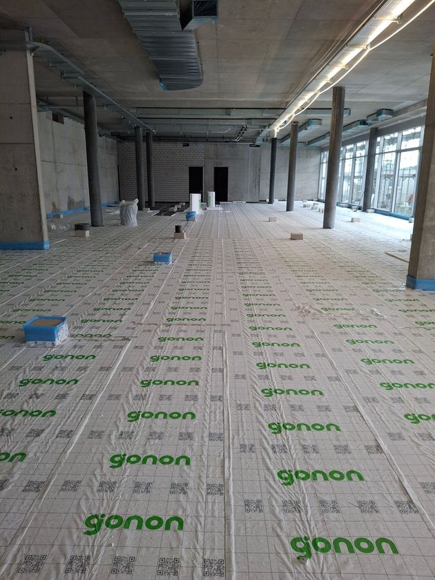 Eine Person in Schutzkleidung installiert Dämmmaterial zwischen den Bodenbalken in einem Raum mit unverputzten Wänden.