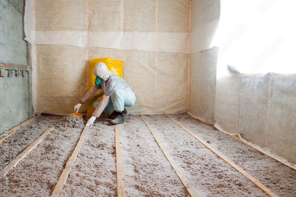 Eine Person in Schutzkleidung installiert Dämmmaterial zwischen den Bodenbalken in einem Raum mit unverputzten Wänden.