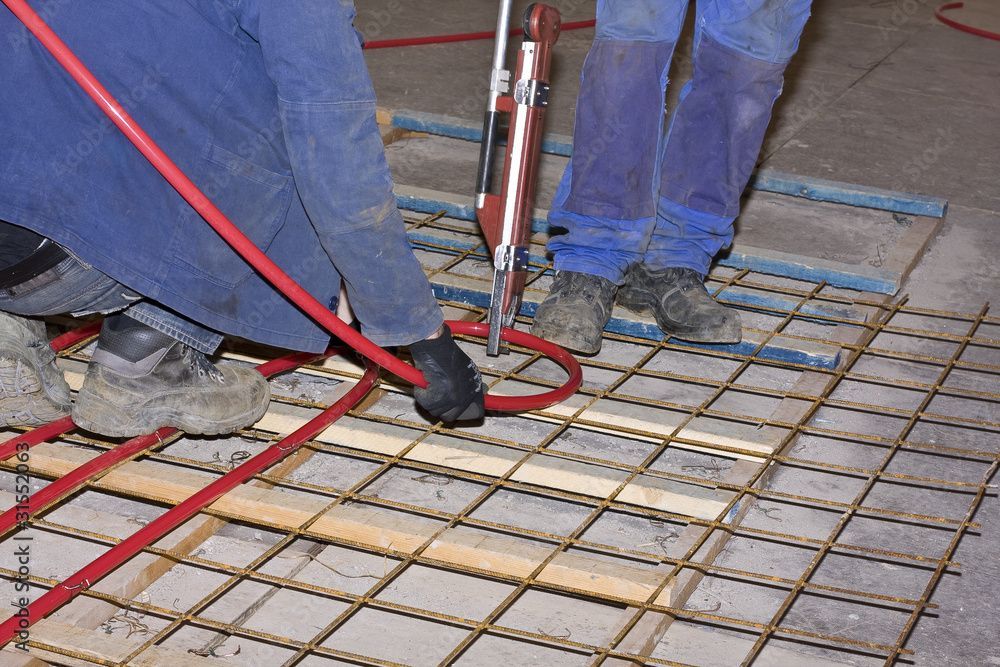 Zwei Personen in Arbeitskleidung montieren rote Heizungsrohre auf einem Metallgittergestell auf einem Betonboden.