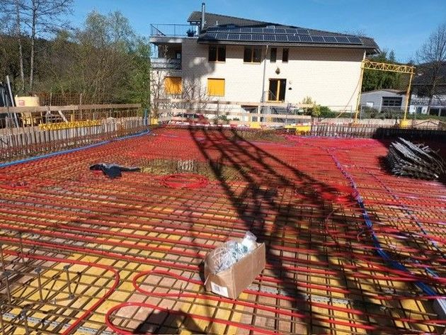 Zwei Personen in Arbeitskleidung montieren rote Heizungsrohre auf einem Metallgittergestell auf einem Betonboden.