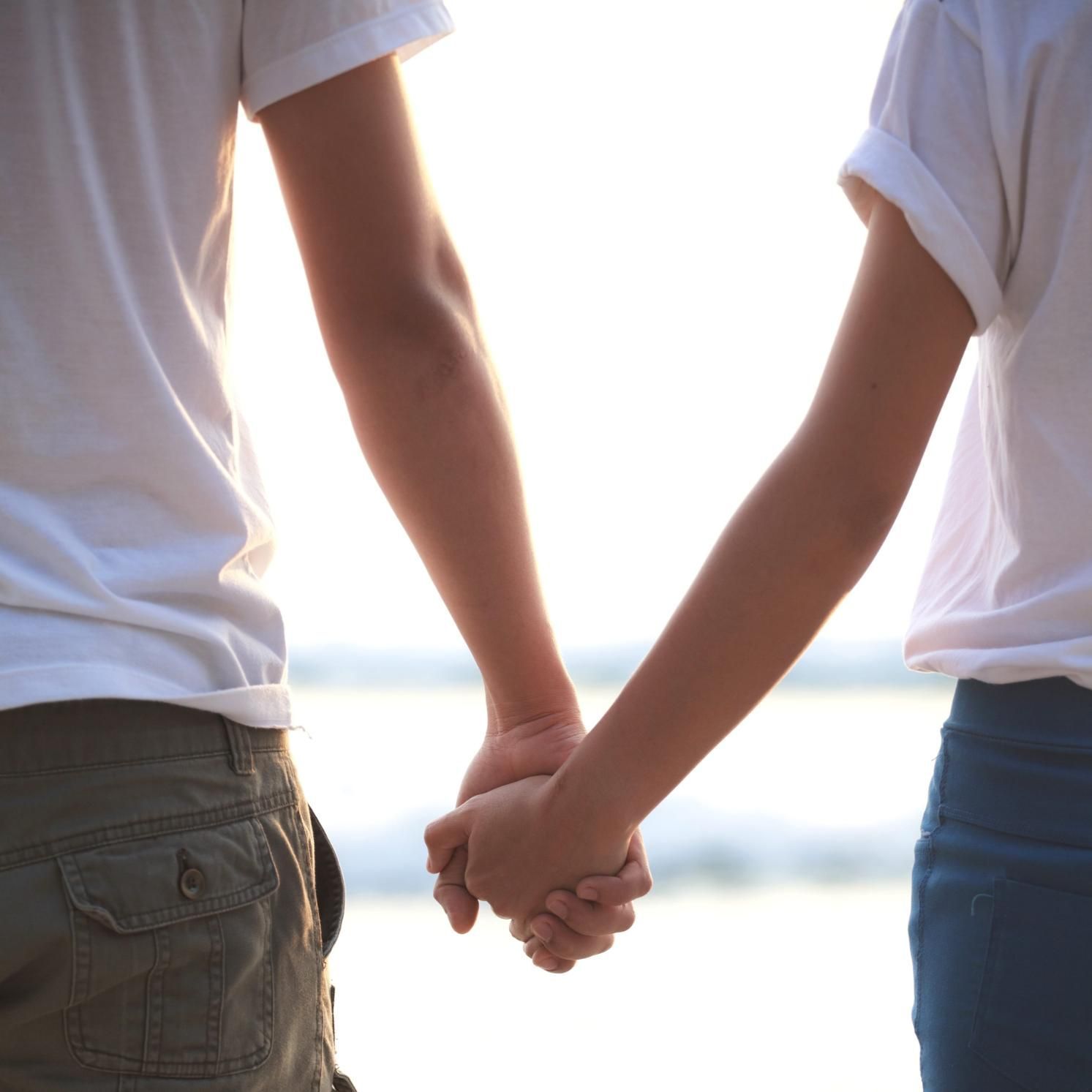 Dos personas tomadas de la mano junto al agua, a contraluz, vestidas con camisas blancas y vaqueros.