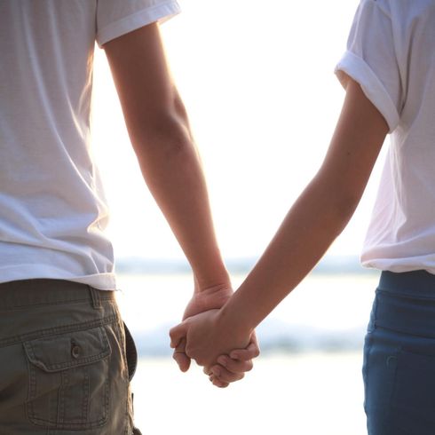 Dos personas tomadas de la mano junto al agua, a contraluz, vestidas con camisas blancas y vaqueros.