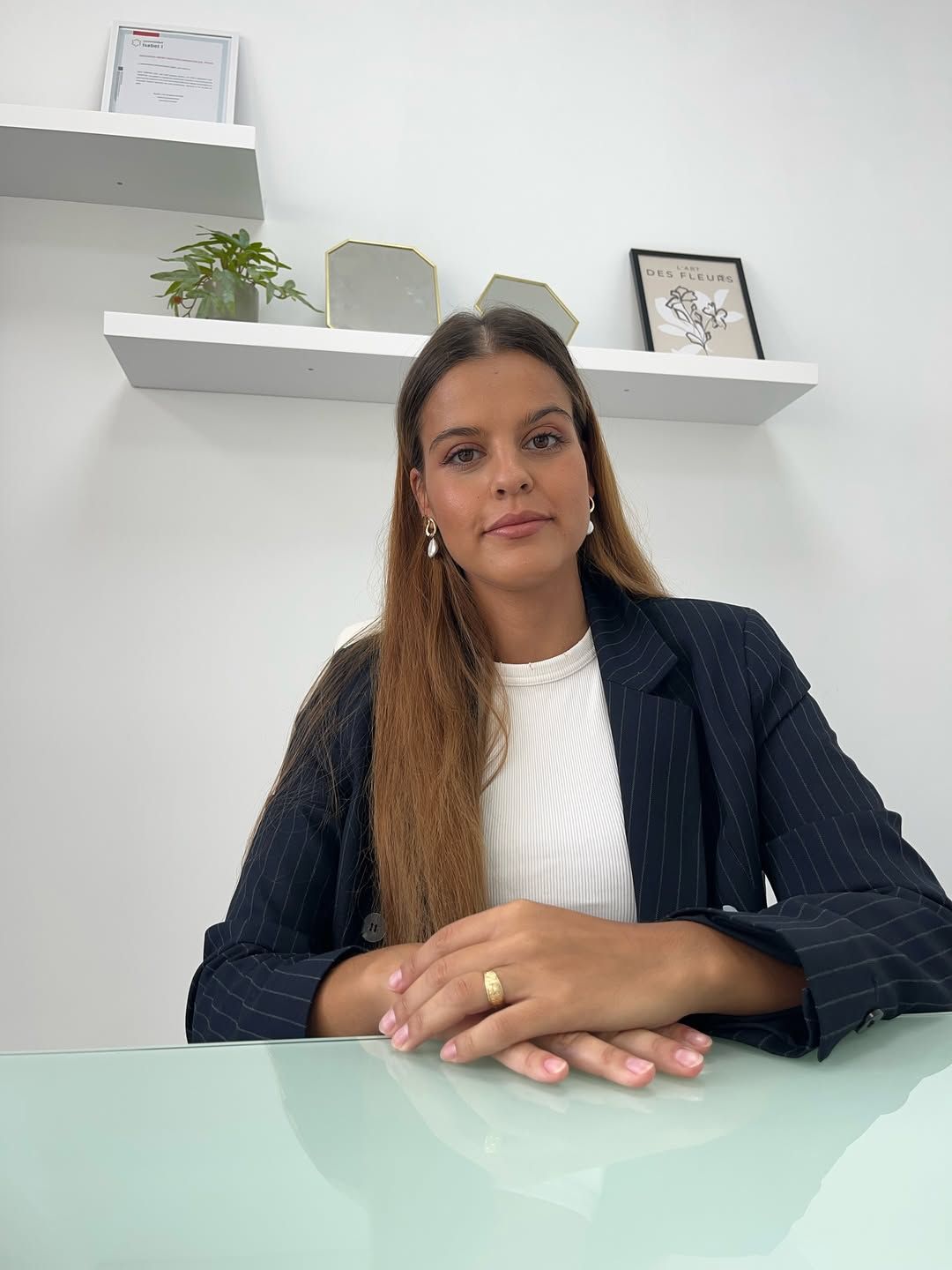 Mujer sentada en un escritorio de cristal en una oficina luminosa, vestida con un blazer oscuro y una blusa blanca.