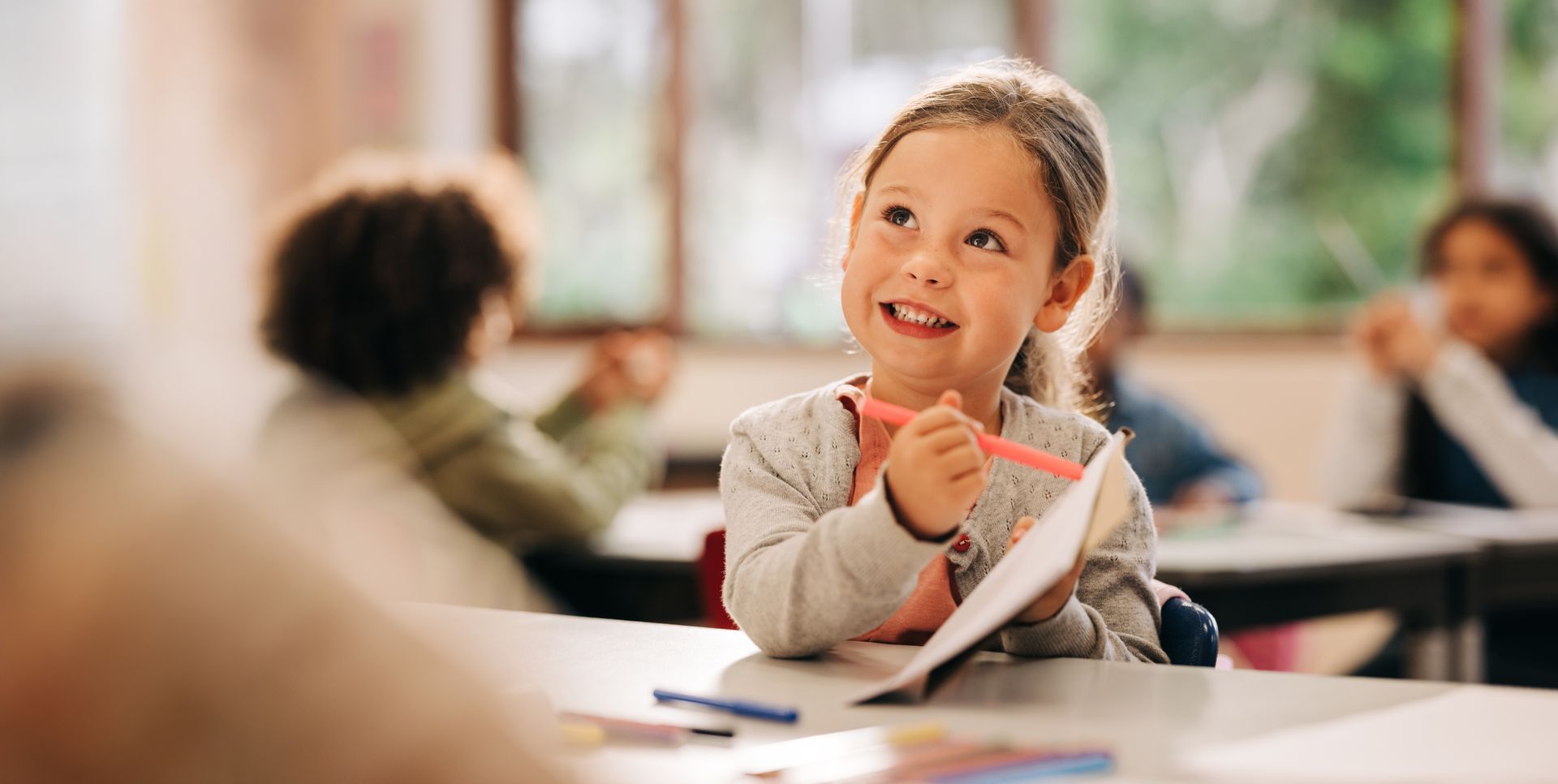 Une petite fille souriante pointe son cahier à l'aide d'un feutre rose