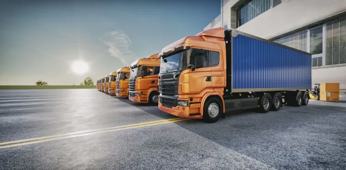 Camiones semirremolque de color naranja estacionados en fila sobre el asfalto, al lado de un edificio, bajo un cielo azul brillante.