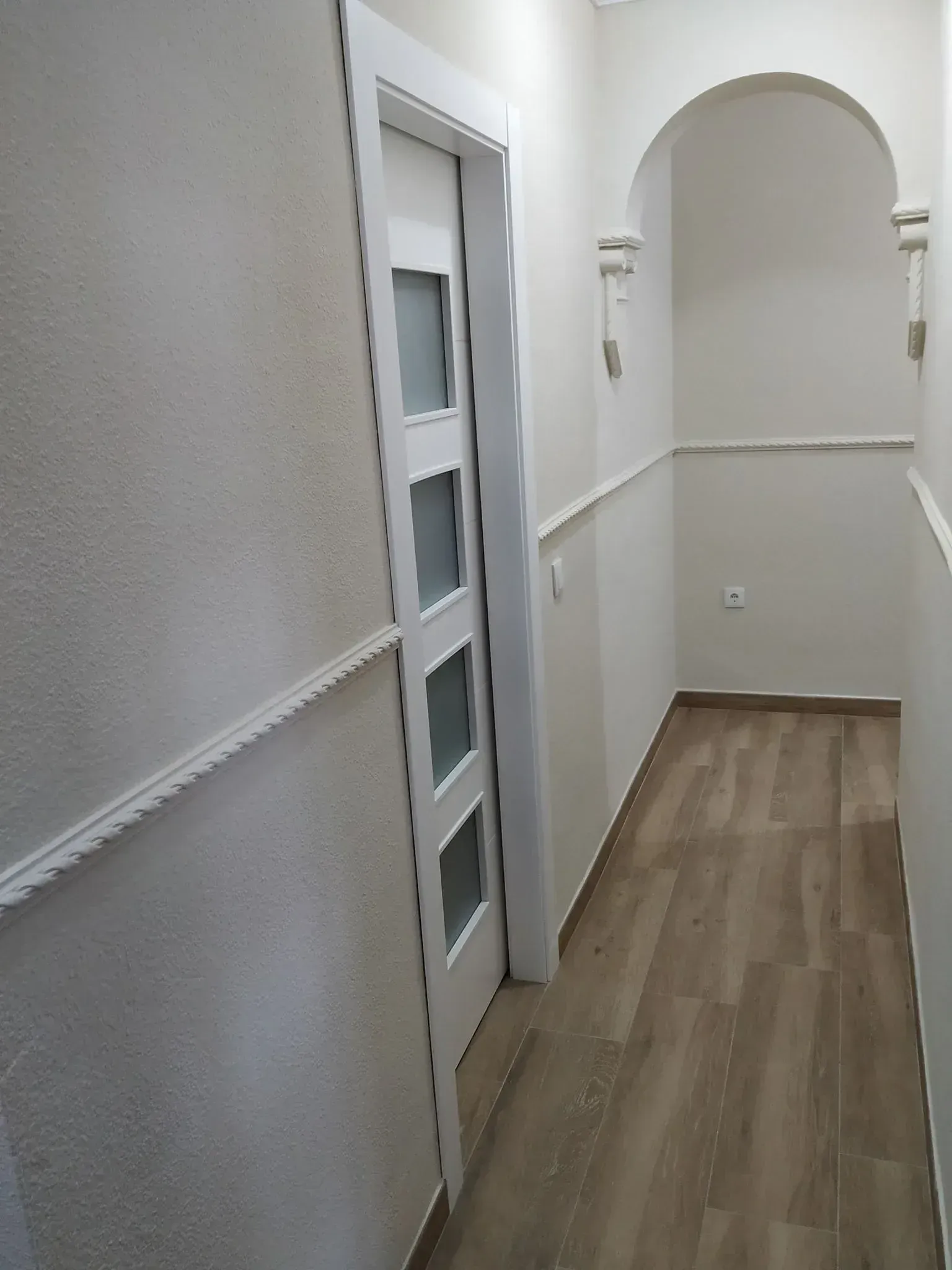 Pasillo estrecho de color blanco con suelo de madera, molduras en las paredes, una puerta a la izquierda y una abertura arqueada al frente.