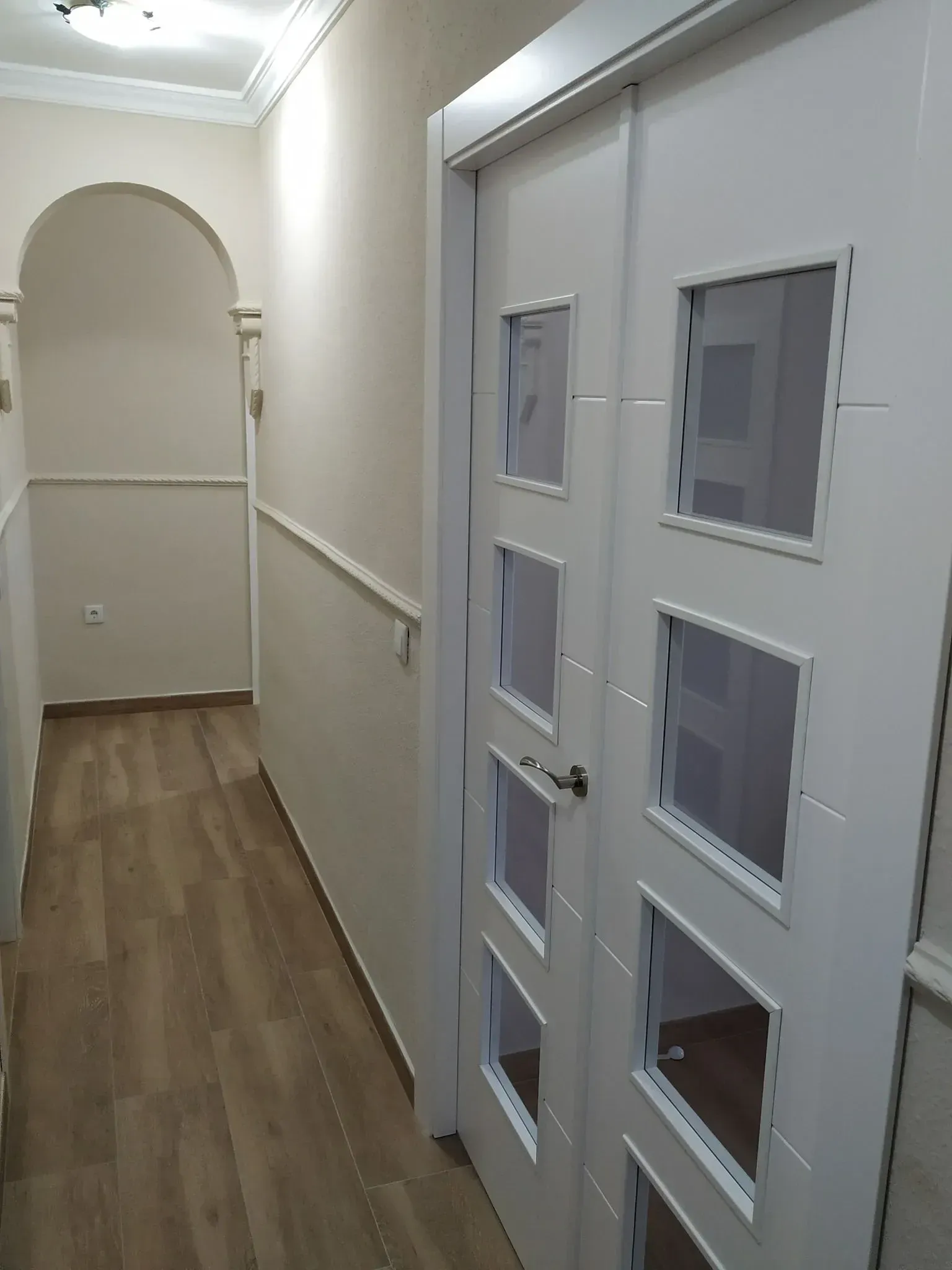 Pasillo estrecho con suelo de madera y paredes blancas, con una puerta blanca y pequeñas ventanas a la derecha.