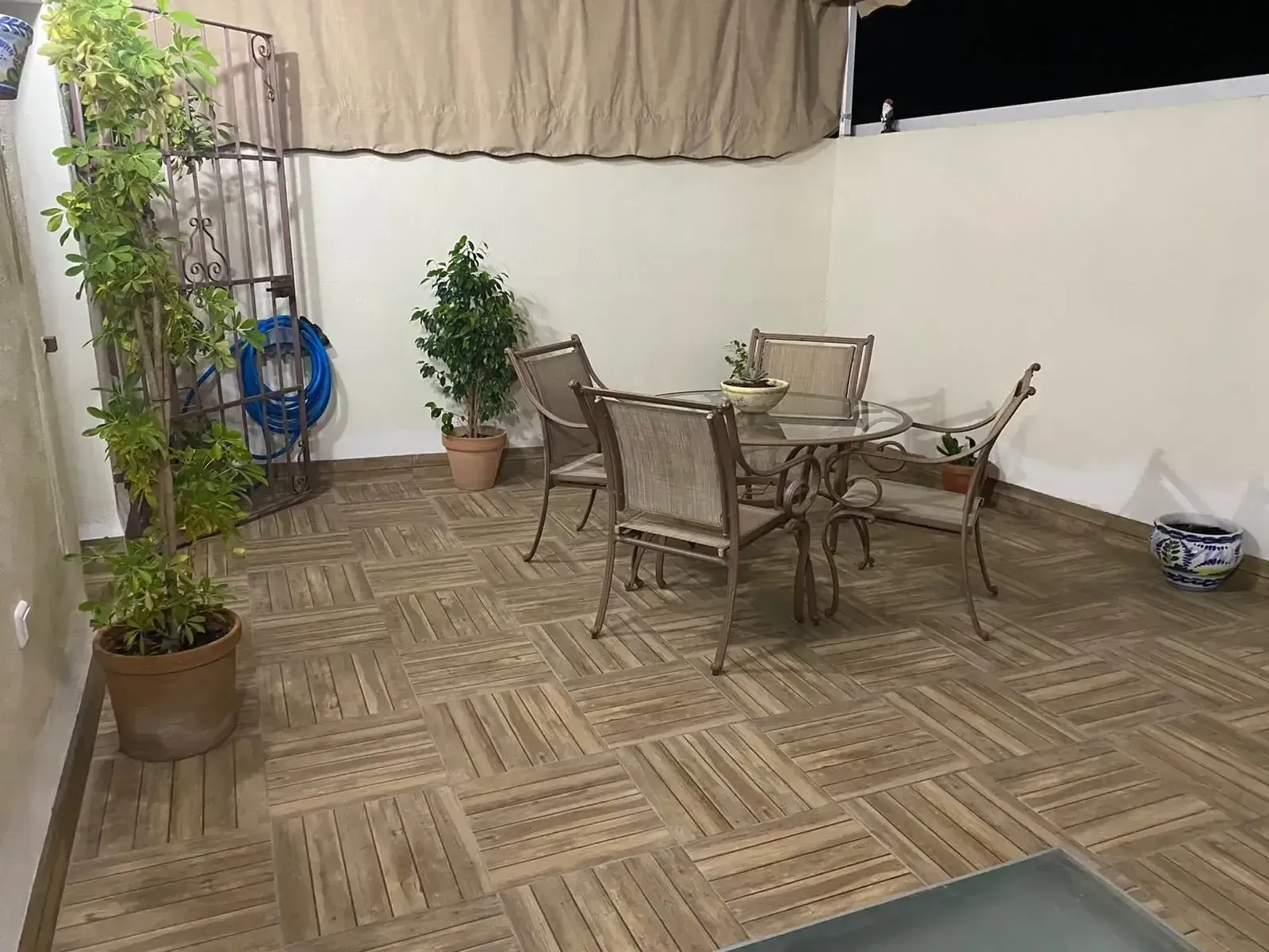 Patio exterior con sillas de mimbre, mesa de cristal, plantas en macetas y suelo de madera con motivos decorativos bajo un toldo.