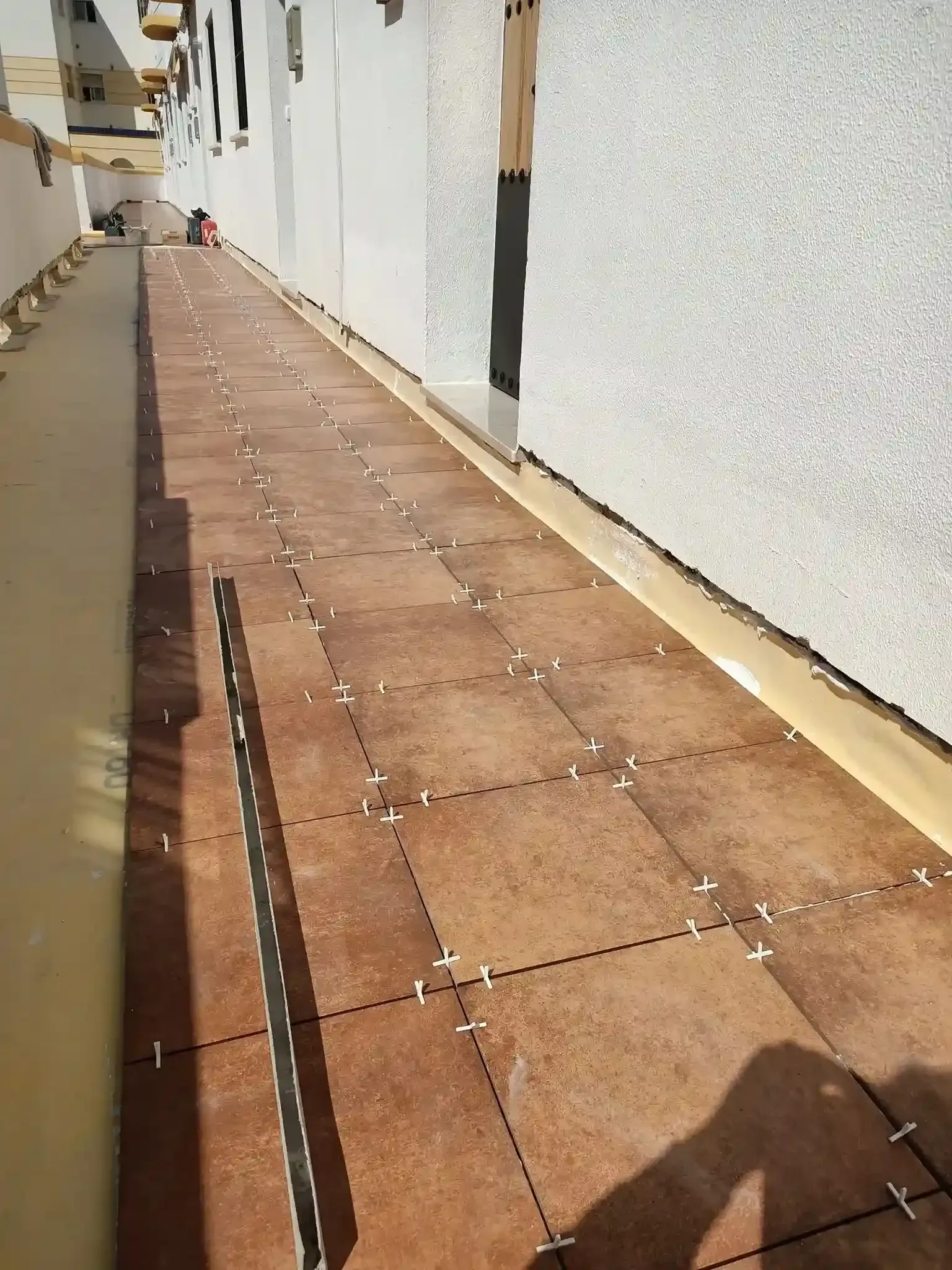 Estrecho pasillo exterior con baldosas marrones junto a un edificio blanco, visto desde un ángulo bajo.