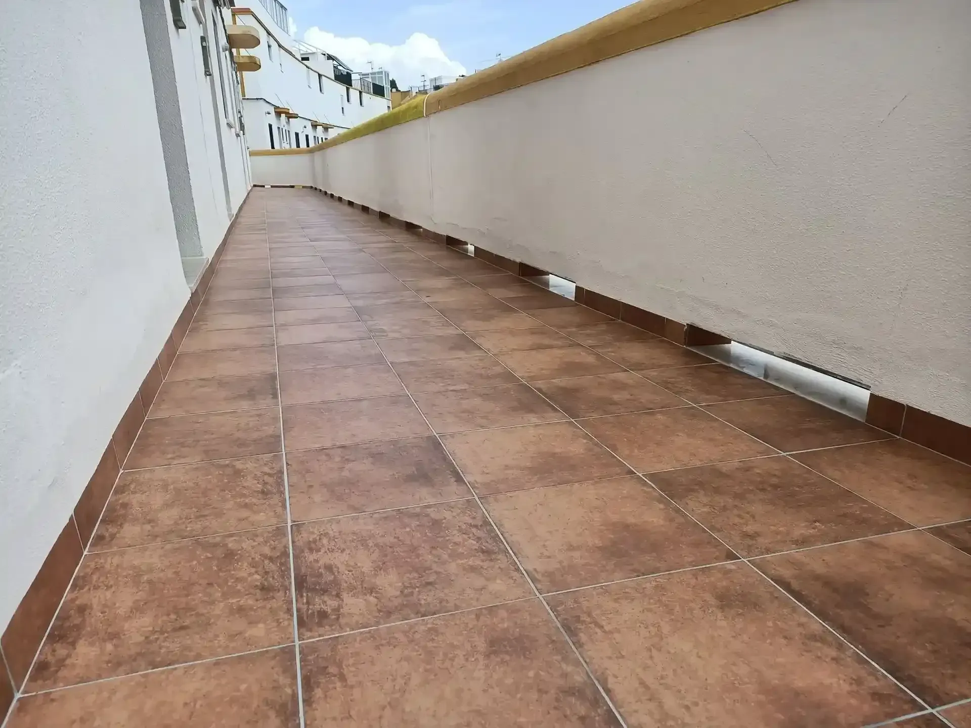 Un largo pasillo exterior embaldosado junto a paredes blancas y una barandilla beige, que se pierde en la distancia.