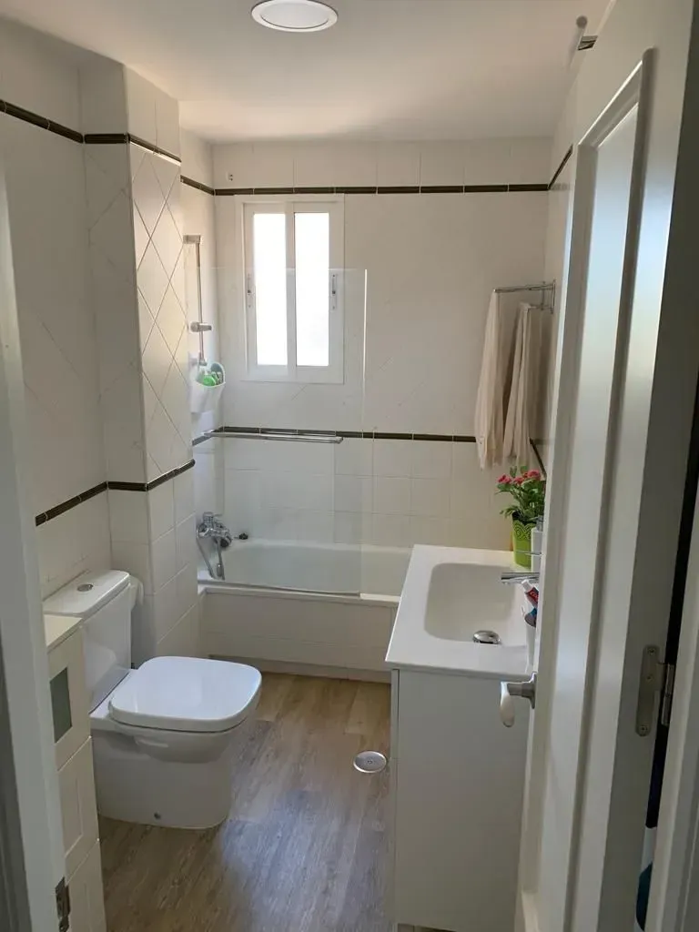 Baño pequeño y moderno con paredes blancas, inodoro, lavabo, bañera y suelo con aspecto de madera.