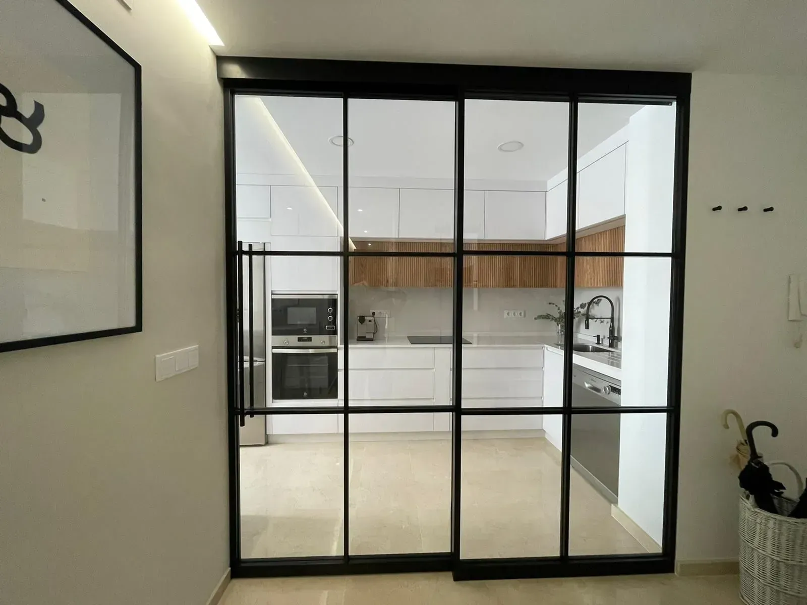 Puertas de cristal dan acceso a una moderna cocina blanca con armarios de madera y una gran isla.