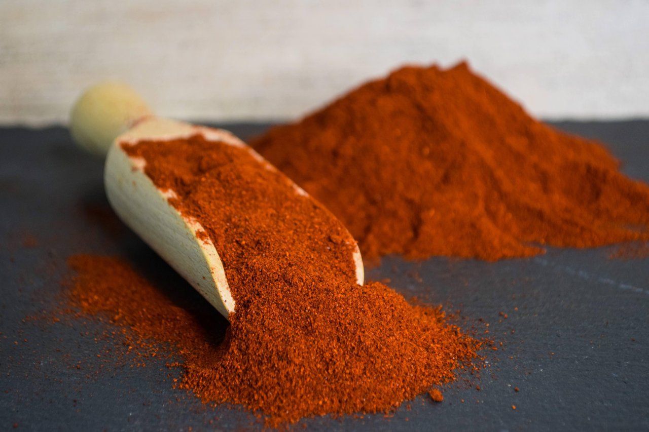Eine Holzschaufel verschüttet leuchtend rotes Gewürzpulver neben einem größeren Haufen auf einer dunklen Oberfläche.
