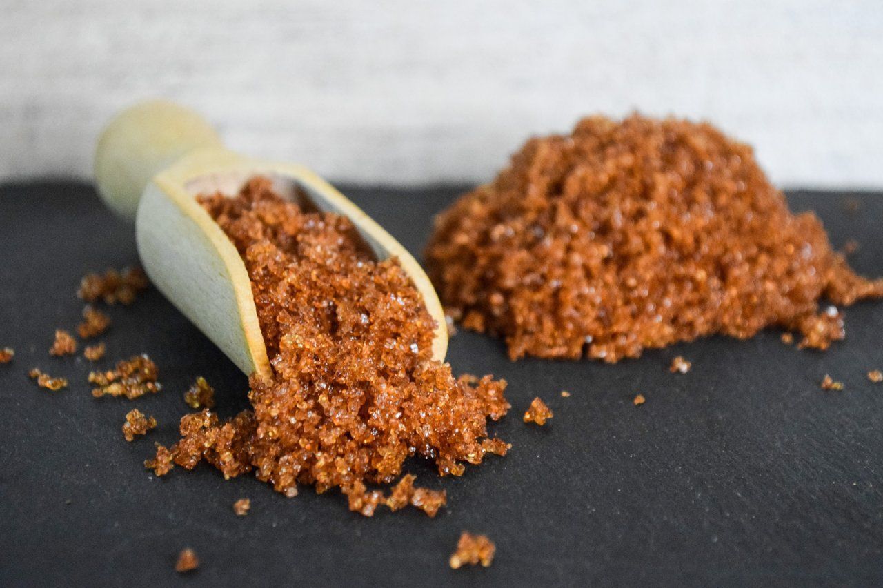 Eine kleine Schaufel verteilt grobes braunes Gewürz oder Würzmittel auf einer dunklen Oberfläche.